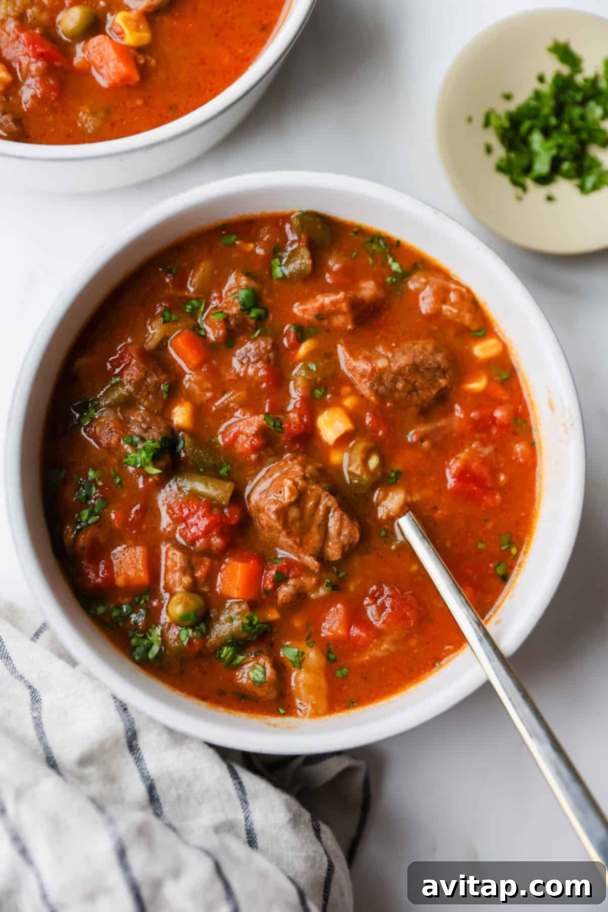 A bowl of beef and vegetable soup with a spoon, featuring chunks of meat, peas, carrots, corn, and tomatoes. A plate of chopped herbs sits nearby on a white surface.