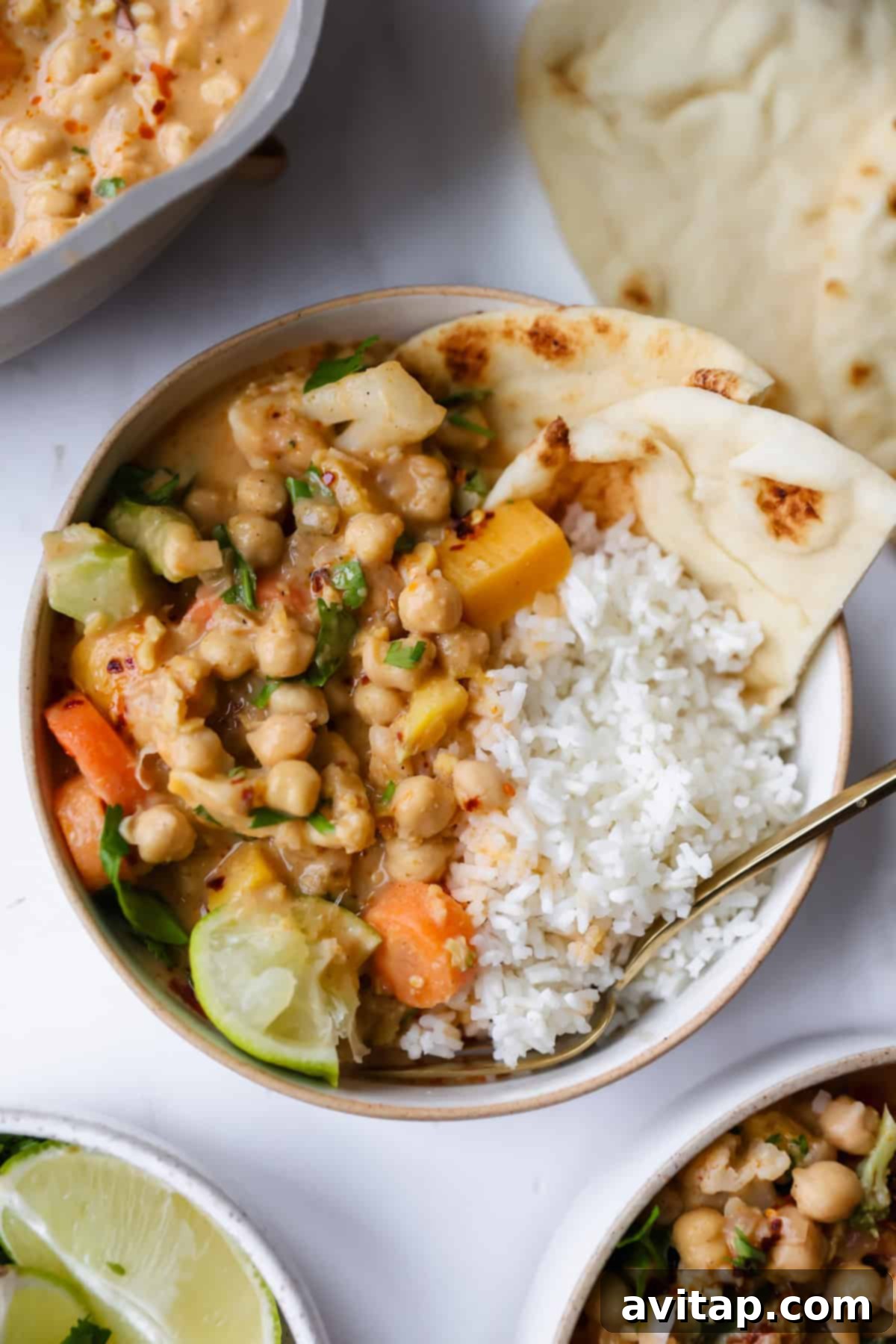 A bowl of chickpea curry with vegetables, white rice, lime wedge, and naan bread, served with a spoon on a white surface.