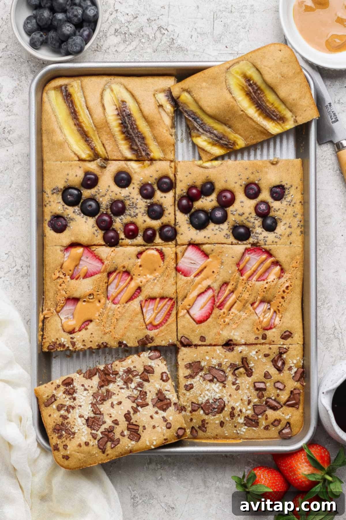Sheet pan protein pancakes with various toppings: banana, blueberries, strawberries with peanut butter, and chocolate chunks.