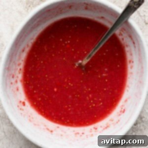 A white bowl filled with red liquid and small seeds, with a metal spoon resting inside, on a light textured surface.