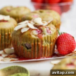Green muffins topped with sliced almonds and bits of strawberry, with a fresh strawberry and a bowl of strawberries in the background; matcha powder in a dish nearby.