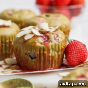 A matcha muffin topped with sliced almonds and strawberry glaze sits next to a whole strawberry, with additional muffins and strawberries in the background.