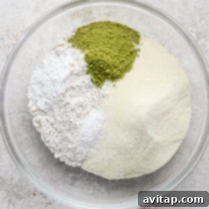 A glass bowl containing four separate dry ingredients: white flour, powdered sugar, green matcha powder, and yellowish almond flour.