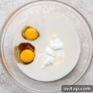 A glass bowl containing two raw eggs, milk, and a dollop of yogurt on a light textured surface.