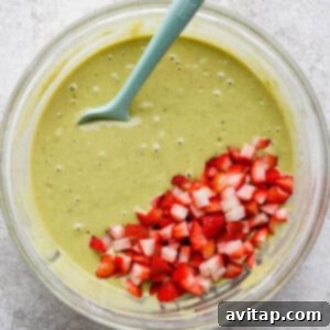A glass bowl filled with green batter and chopped strawberries, with a blue spatula resting inside, placed on a light textured surface.