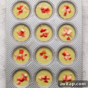 A muffin tin with twelve unbaked cupcake liners filled with batter and topped with chopped strawberries, viewed from above.