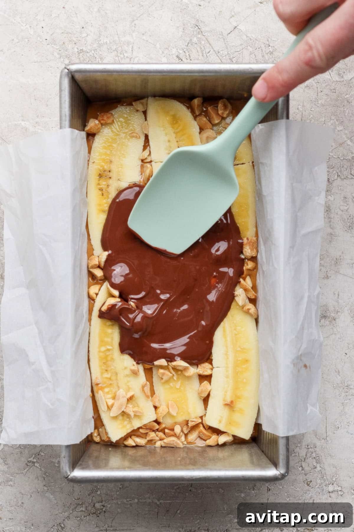 Melted chocolate being spread over sliced bananas and chopped peanuts in a parchment-lined baking pan.