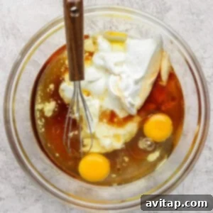 A glass bowl containing two raw eggs, yogurt, and a brown liquid with a metal whisk resting on the side, placed on a textured countertop.
