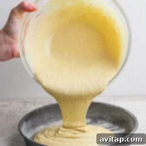A hand pours cake batter from a glass bowl into a round, floured metal baking pan on a light countertop.