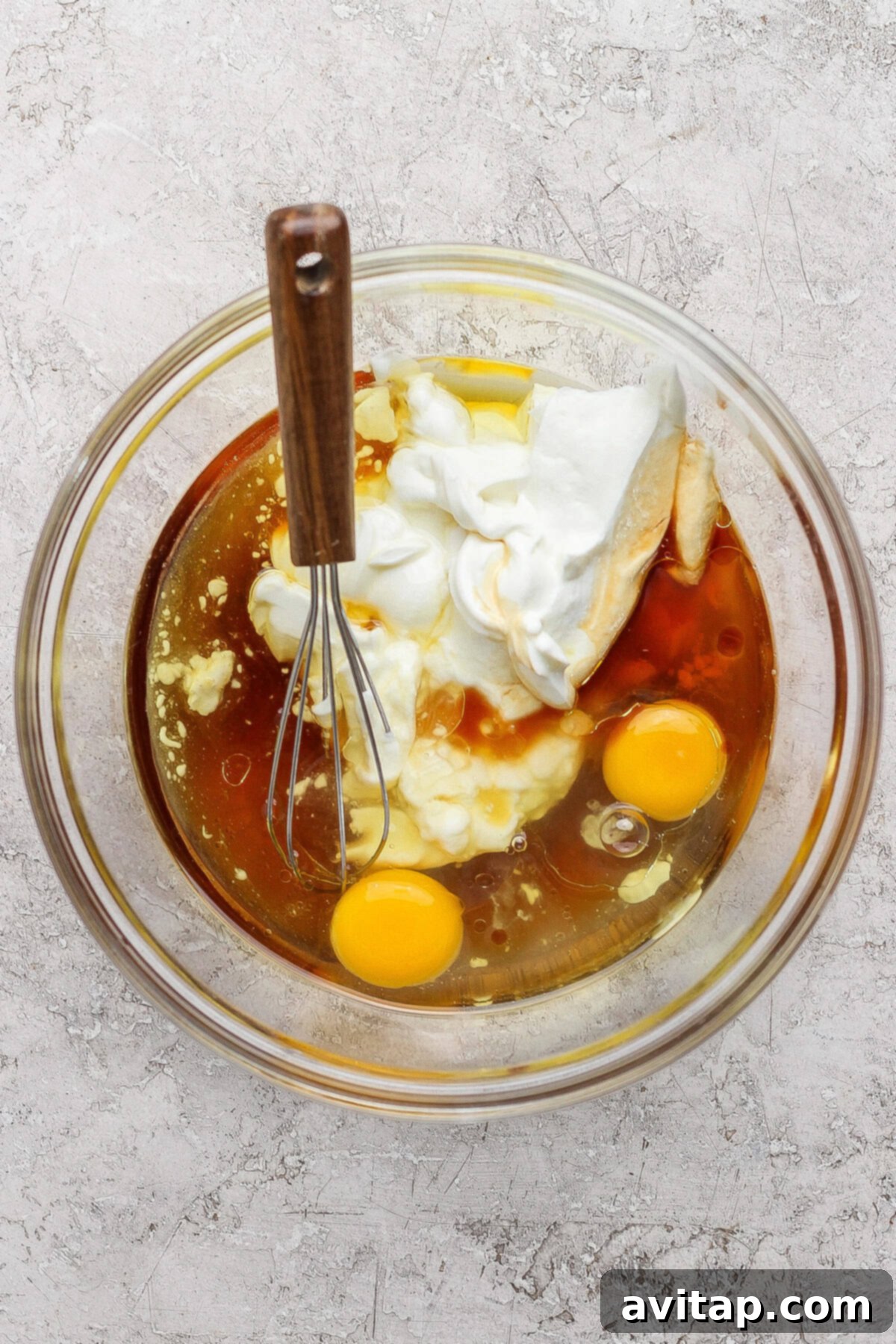 A glass bowl containing two raw eggs, yogurt, and a brown liquid with a metal whisk resting on the side, placed on a textured countertop.