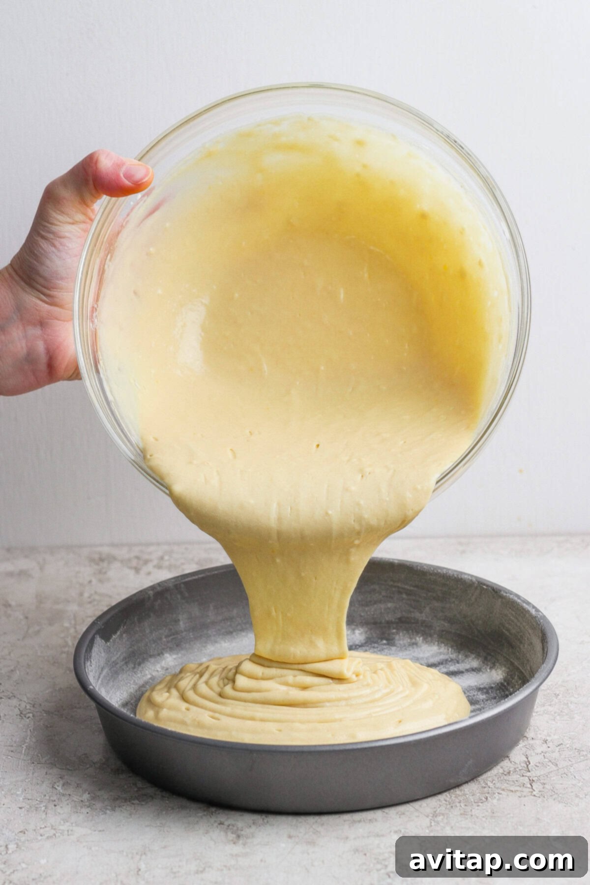 A hand pours cake batter from a glass bowl into a round, floured metal baking pan on a light countertop.