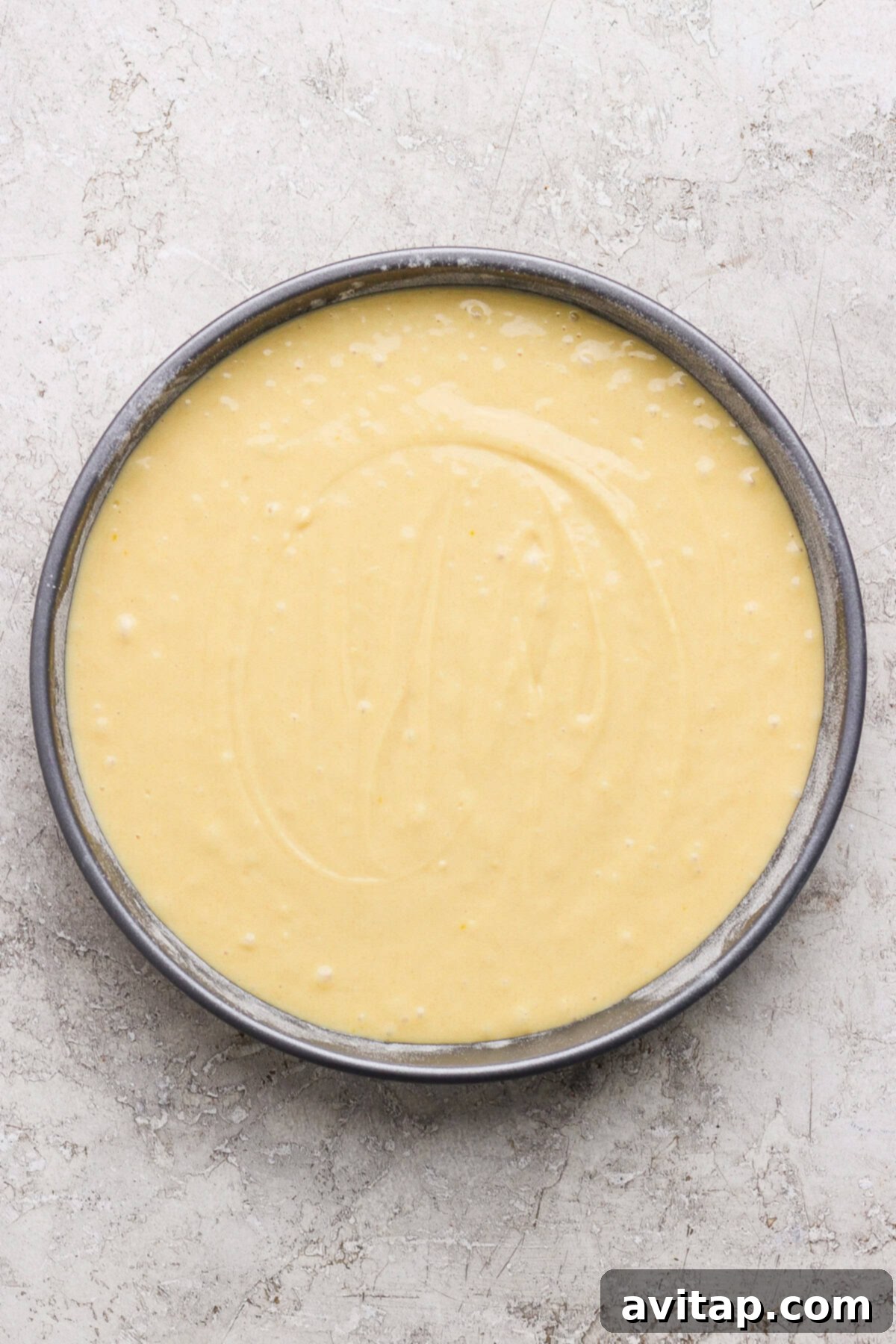 A round cake pan filled with smooth, pale yellow cake batter sits on a light textured surface.