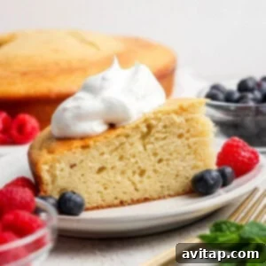 A slice of yellow cake topped with whipped cream, served on a plate with fresh raspberries and blueberries; whole cake and more berries in the background.