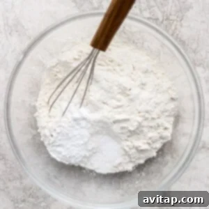 A glass bowl containing flour and baking powder with a metal whisk resting on top, placed on a light textured surface.