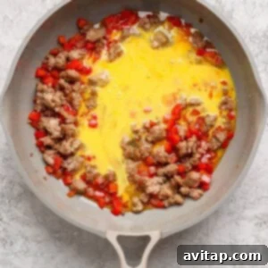 A skillet containing cooked sausage, diced red bell peppers, and beaten eggs beginning to cook.