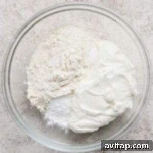 A glass bowl containing flour, baking powder, salt, and plain yogurt on a textured light surface.