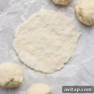 Four dough balls and one flattened round of dough sit on a sheet of parchment paper dusted with flour.
