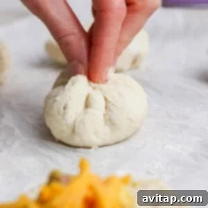 A hand pinches the top of a ball of dough to seal it closed; fillings and shredded cheese are visible on a piece of dough in the foreground.