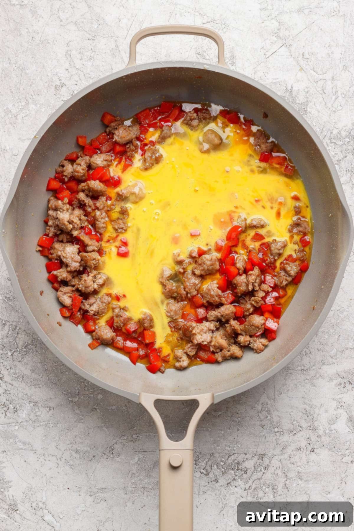 A non-stick skillet on a stovetop, filled with cooked ground sausage, diced red bell peppers, and scrambled eggs that are just beginning to set.