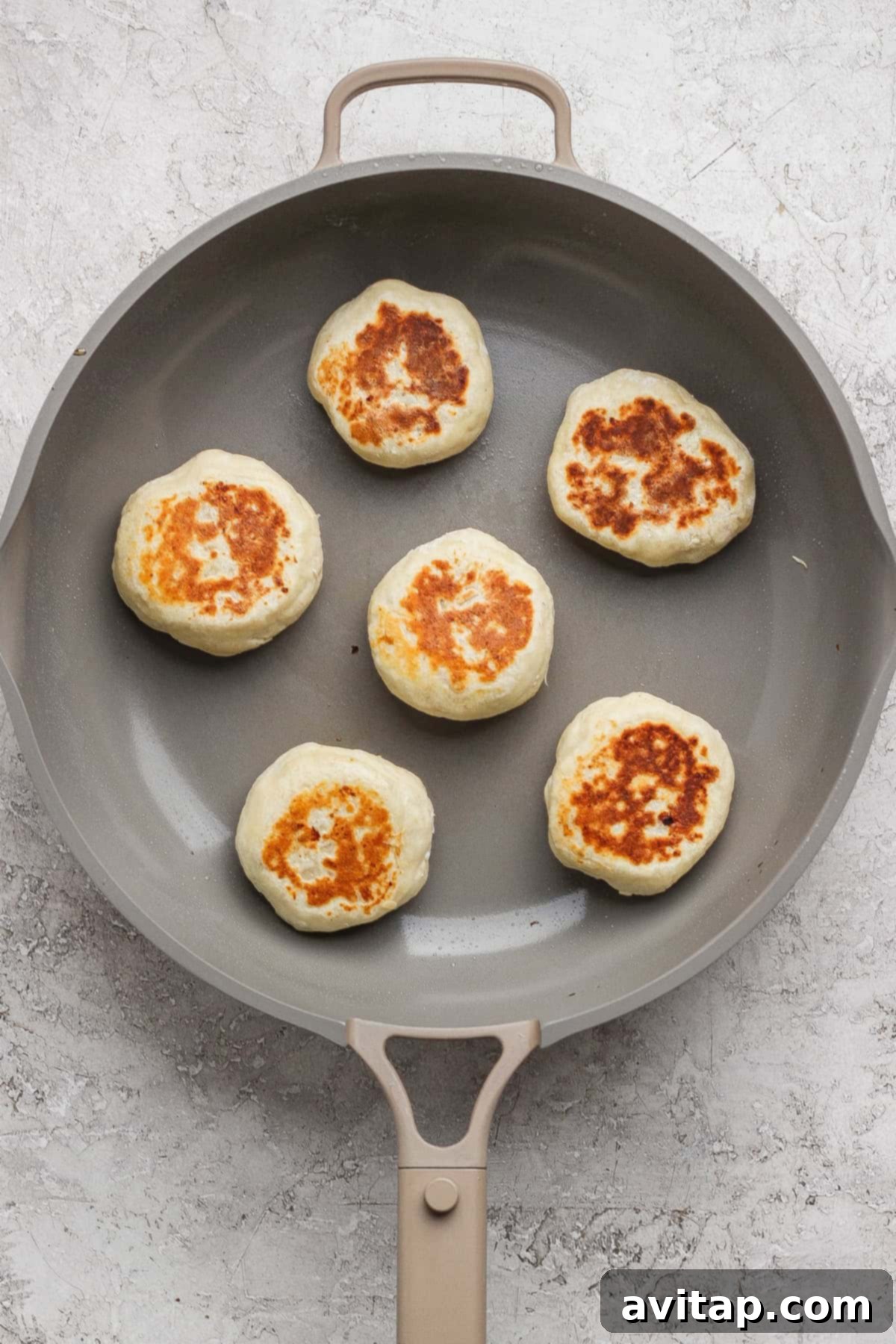 Six round, golden-brown stuffed English muffins sizzling gently in a nonstick skillet on a light kitchen counter.