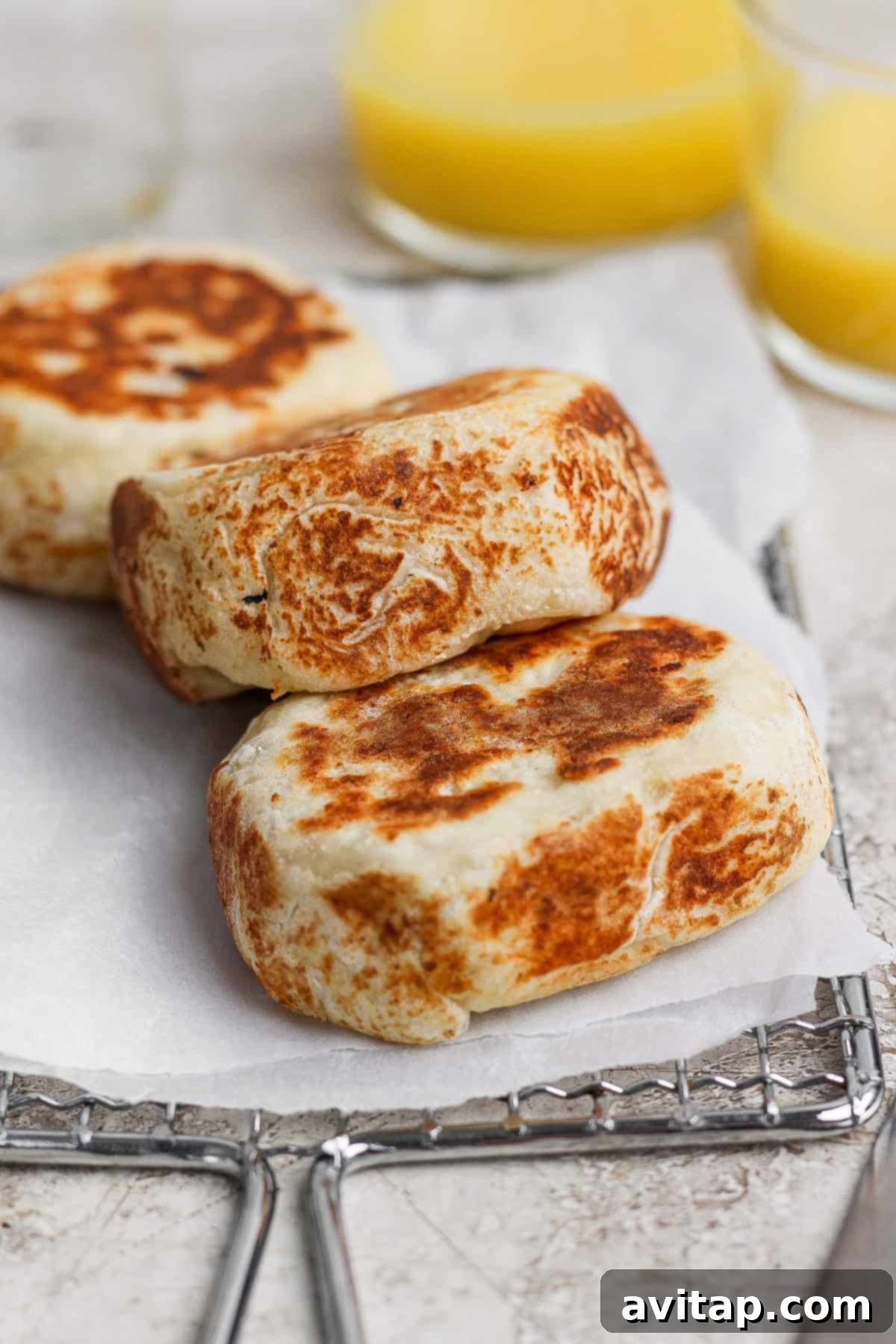 Three perfectly browned, thick stuffed English muffins cooling on parchment paper on a wire rack, with blurred glasses of orange juice in the background, suggesting a complete breakfast.