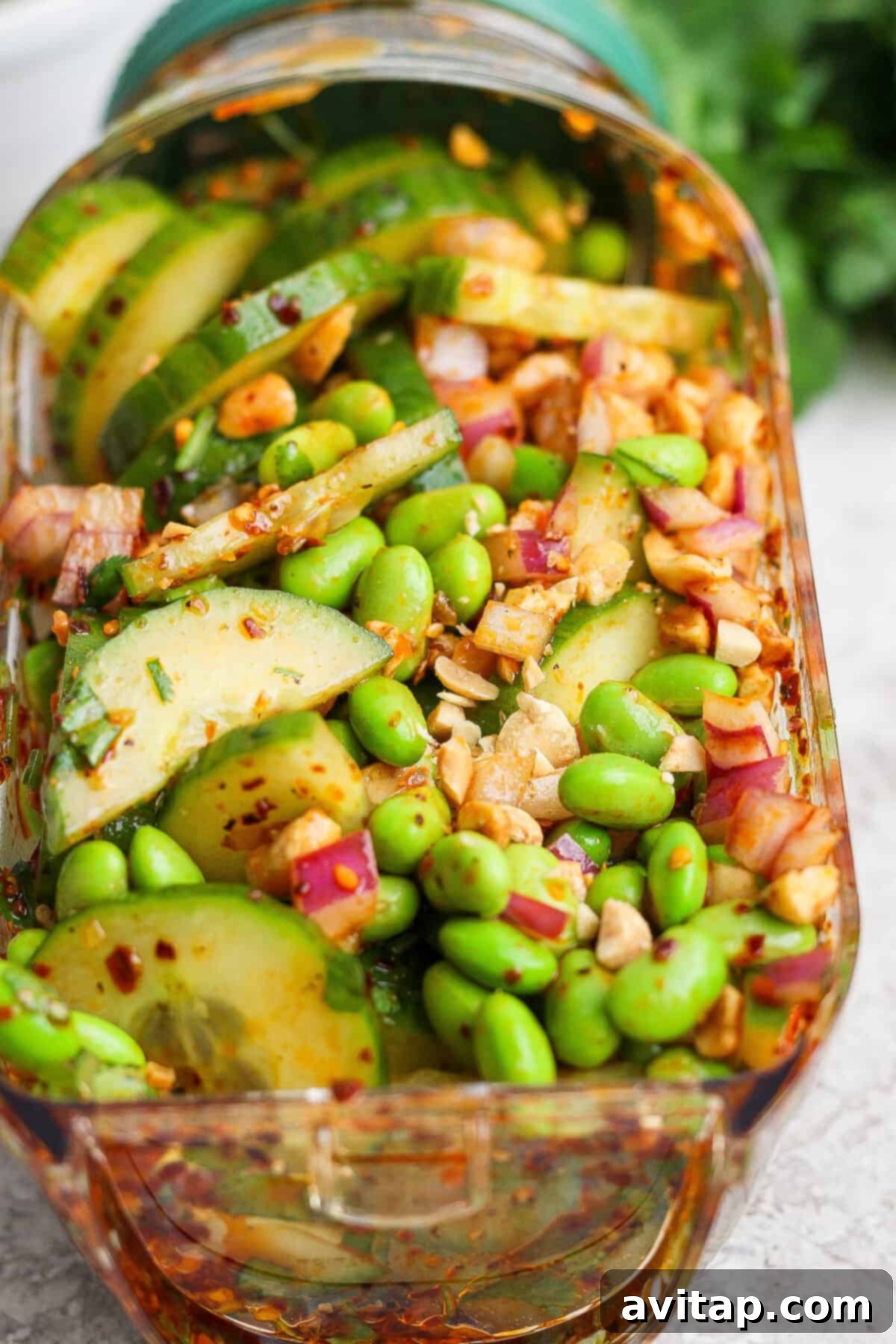 A close-up of a vibrant chili crunch edamame cucumber salad with sliced cucumbers, green edamame, red onion, chopped nuts, and a bright red chili-flake dressing, presented in a clear rectangular glass container.