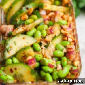 A close-up of a vibrant chili crunch edamame cucumber salad with sliced cucumbers, green edamame, red onion, chopped nuts, and a bright red chili-flake dressing, presented in a clear rectangular glass container.