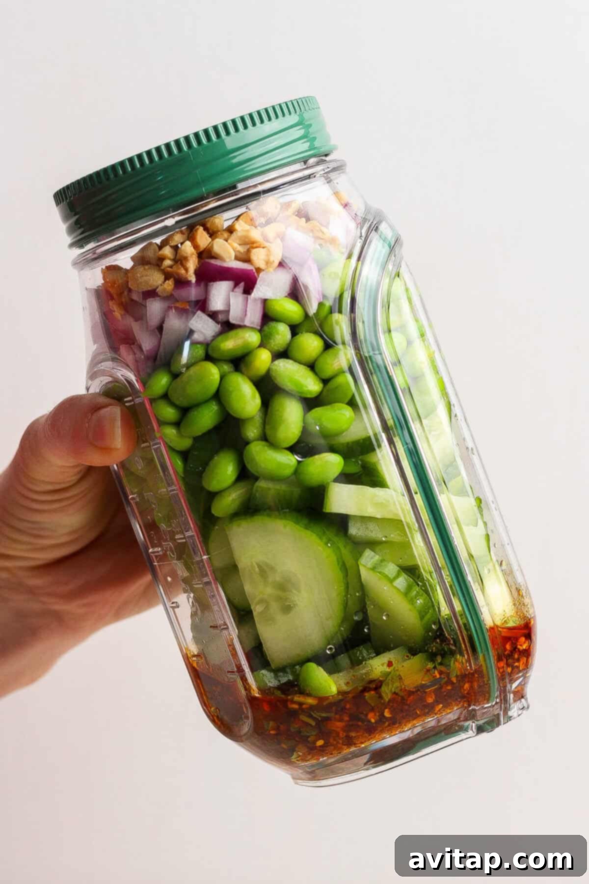 A hand holds a tilted plastic jar with layers of chili crunch edamame cucumber salad ingredients: dressing at the bottom, then cucumber, edamame, red onion, and chopped peanuts.