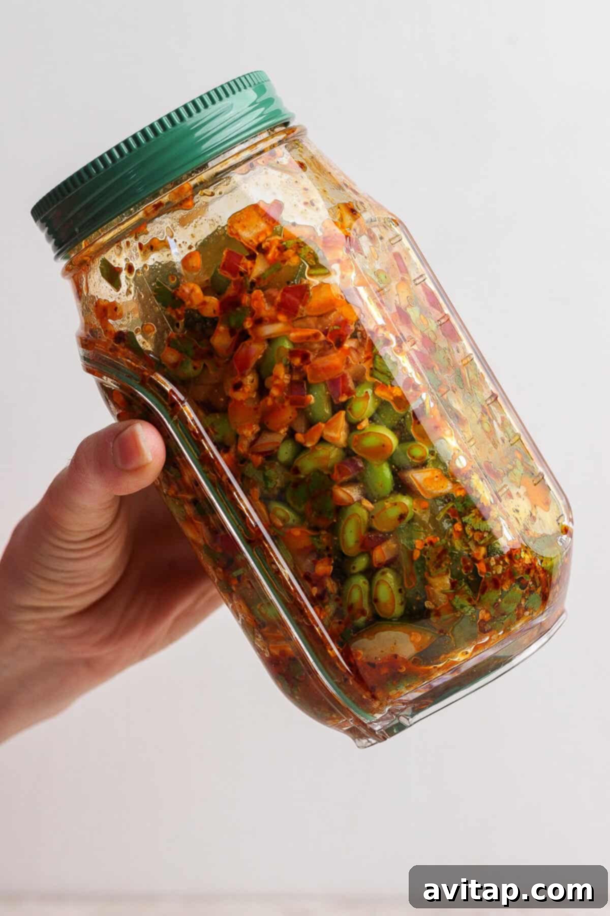 A hand holds a glass jar filled with colorful, chopped vegetables and spices, including edamame, cucumber, and red onion, with a green lid, ready for serving.