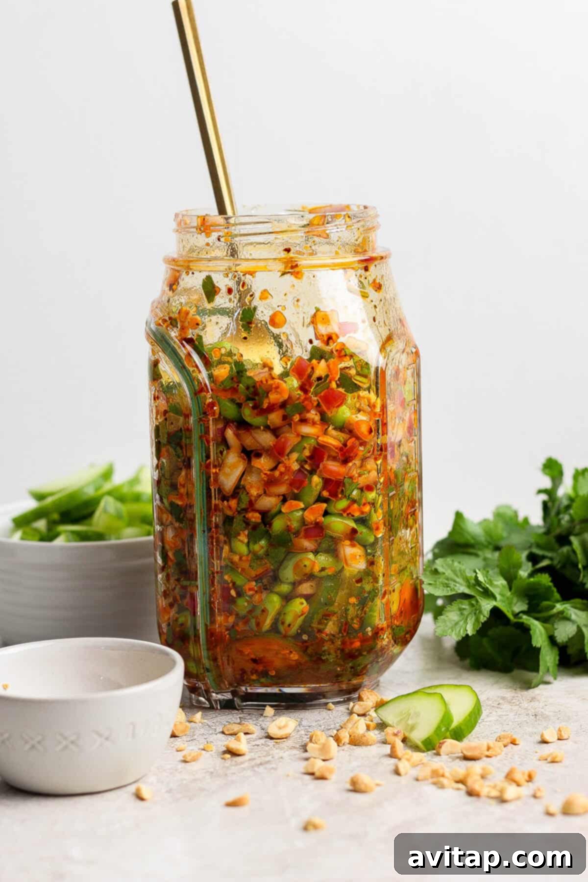 A glass jar filled with a vibrant chili crunch edamame cucumber salad, topped with dressing and a spoon, sits on a countertop beside fresh herbs, peanuts, and cucumber slices.