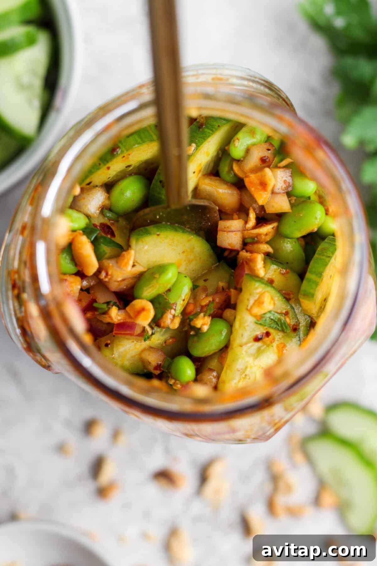 A jar filled with a colorful mix of sliced cucumbers, edamame, chopped red onions, and peanuts, seasoned with chili crunch dressing. A spoon is inside, and fresh ingredients are scattered around on a light surface.