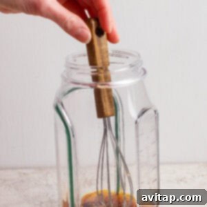 A hand mixing chili crunch dressing ingredients with a whisk in a clear glass jar on a light surface.