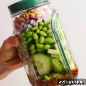 A hand adding layers of cucumber, edamame, red onion, and walnuts to a tilted plastic jar with dressing already at the bottom, preparing a layered chili crunch salad.