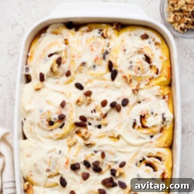 A baking dish brimming with freshly baked carrot cake cinnamon rolls, generously frosted and garnished with a sprinkling of golden raisins and crunchy walnuts. A small bowl of extra walnuts sits nearby, ready for topping.