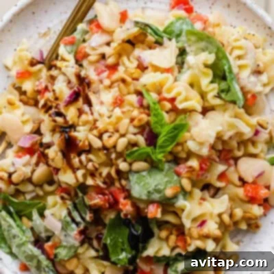 A plate of pasta salad with spinach, pine nuts, white beans, chopped red onions, red bell peppers, and a balsamic drizzle, garnished with fresh basil.