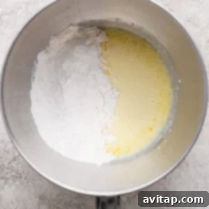 A metal mixing bowl containing flour on one side and a yellow liquid mixture on the other, placed on a light textured surface.