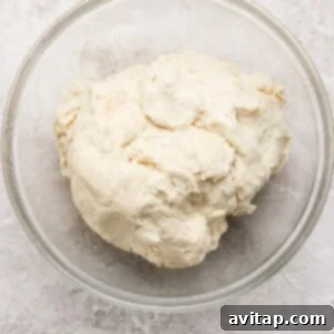 A clear glass bowl containing a ball of raw, unbaked dough sits on a light textured surface.