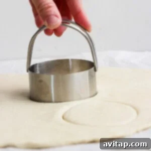 A hand presses a round metal biscuit cutter into rolled-out dough on a flat surface.