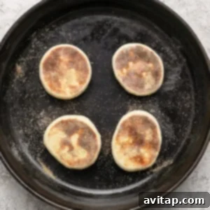 Four partially cooked English muffins in a cast iron skillet on a light textured surface.