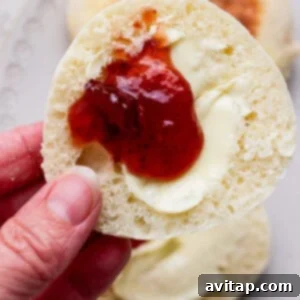 A hand holds half of an English muffin topped with melted butter and strawberry jam, with more muffins on a plate in the background.