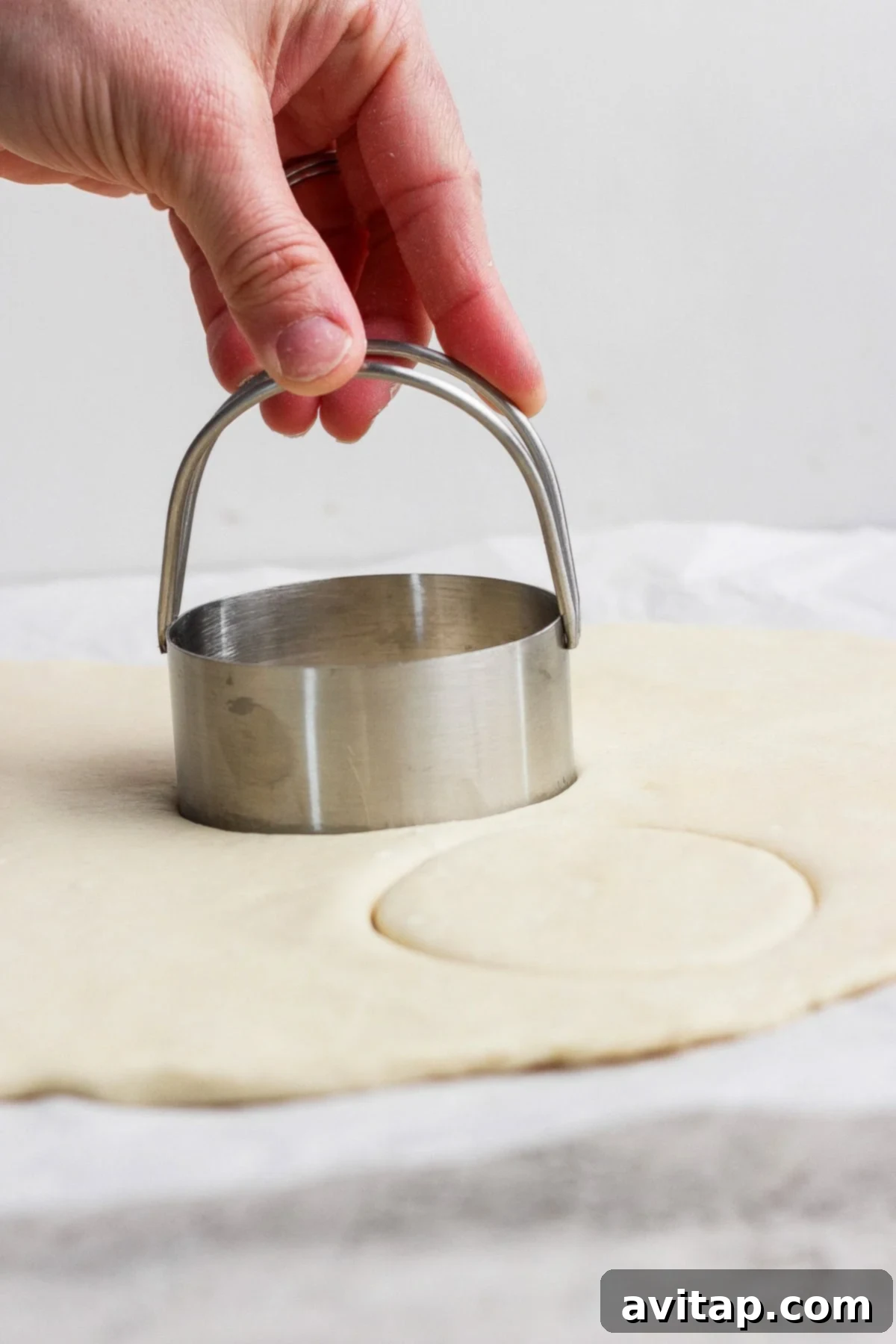 A hand presses a round metal biscuit cutter into rolled-out dough on a flat surface.