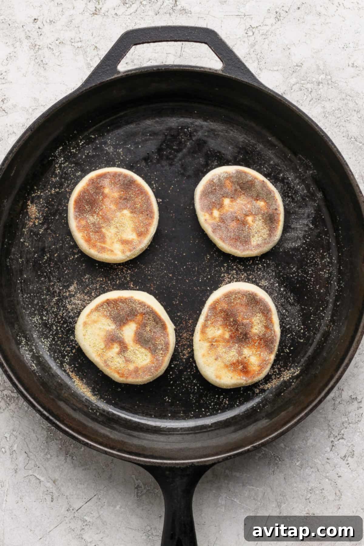 Four partially cooked English muffins in a cast iron skillet on a light textured surface.