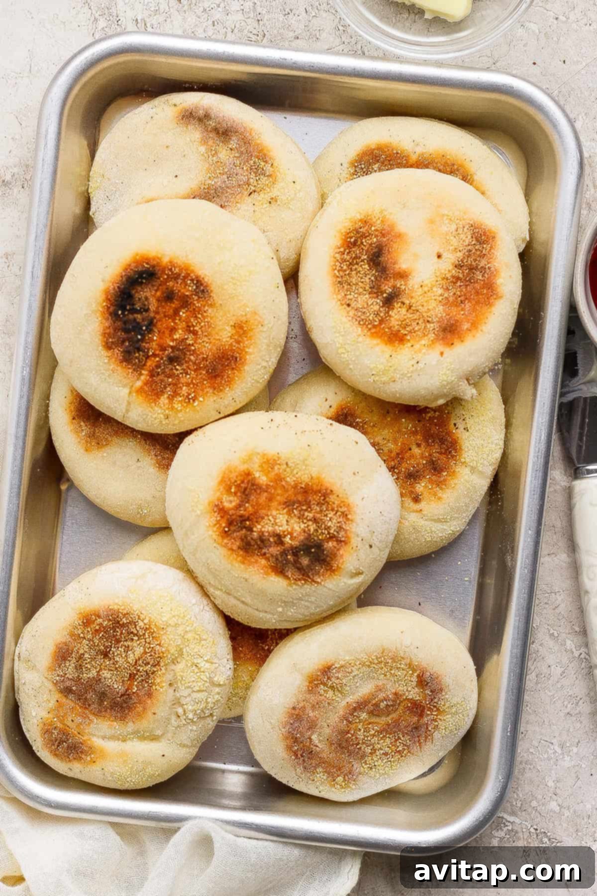 A metal tray filled with nine homemade English muffins, each with a golden-brown, toasted top and a light dusting of cornmeal.