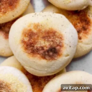 A close-up of several golden-brown English muffins topped with cornmeal, stacked together on a light surface.