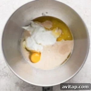 A mixing bowl containing yogurt, one egg, melted butter, and liquid yeast mixture on a light textured surface.
