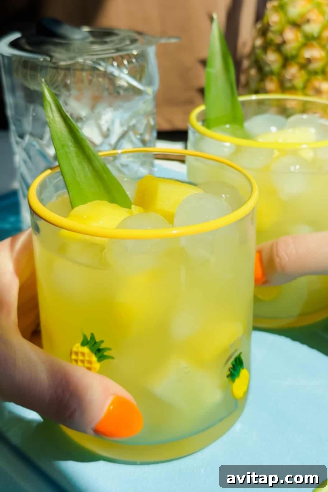 Two hands holding glasses filled with ice, yellow drink, pineapple chunks, and a pineapple leaf garnish, decorated with small pineapple designs.