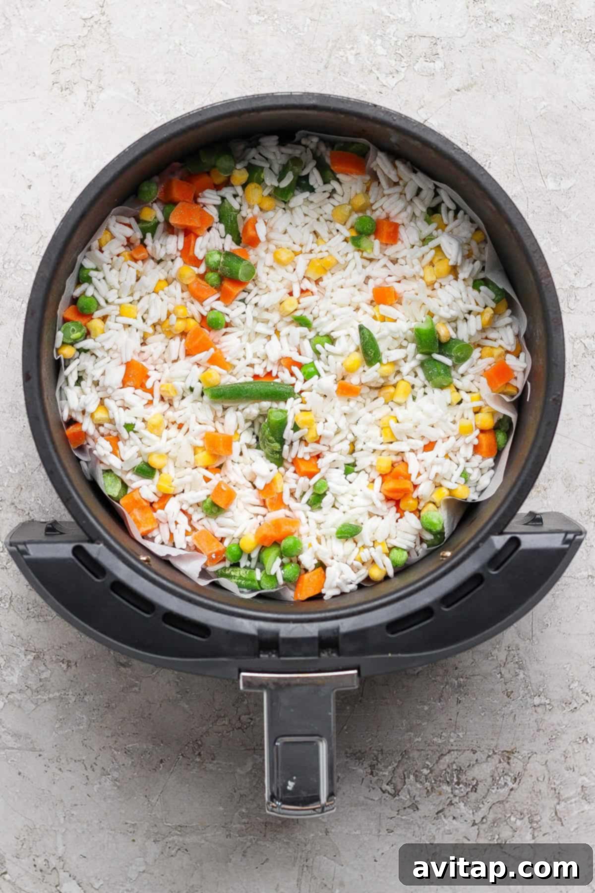 An air fryer basket filled with uncooked white rice and mixed vegetables including carrots, peas, corn, and green beans.