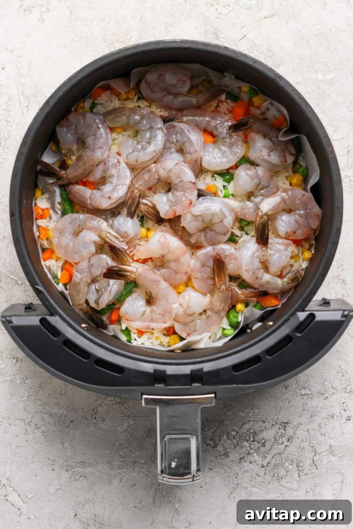 Raw shrimp arranged on top of mixed vegetables and rice in the basket of an air fryer.