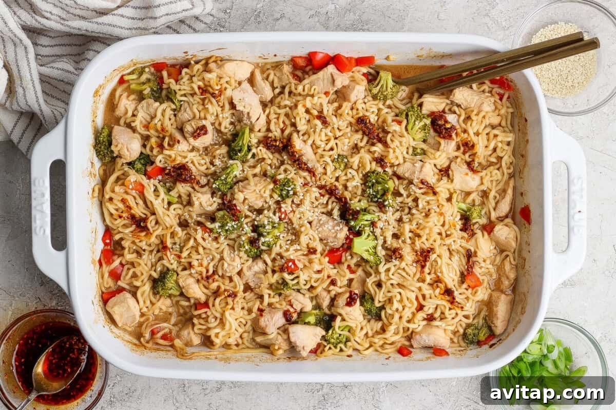 Rectangular white baking dish filled with cooked ramen noodles, chicken pieces, broccoli, and red peppers, garnished with sesame seeds and sauce, with chopsticks resting on the dish.