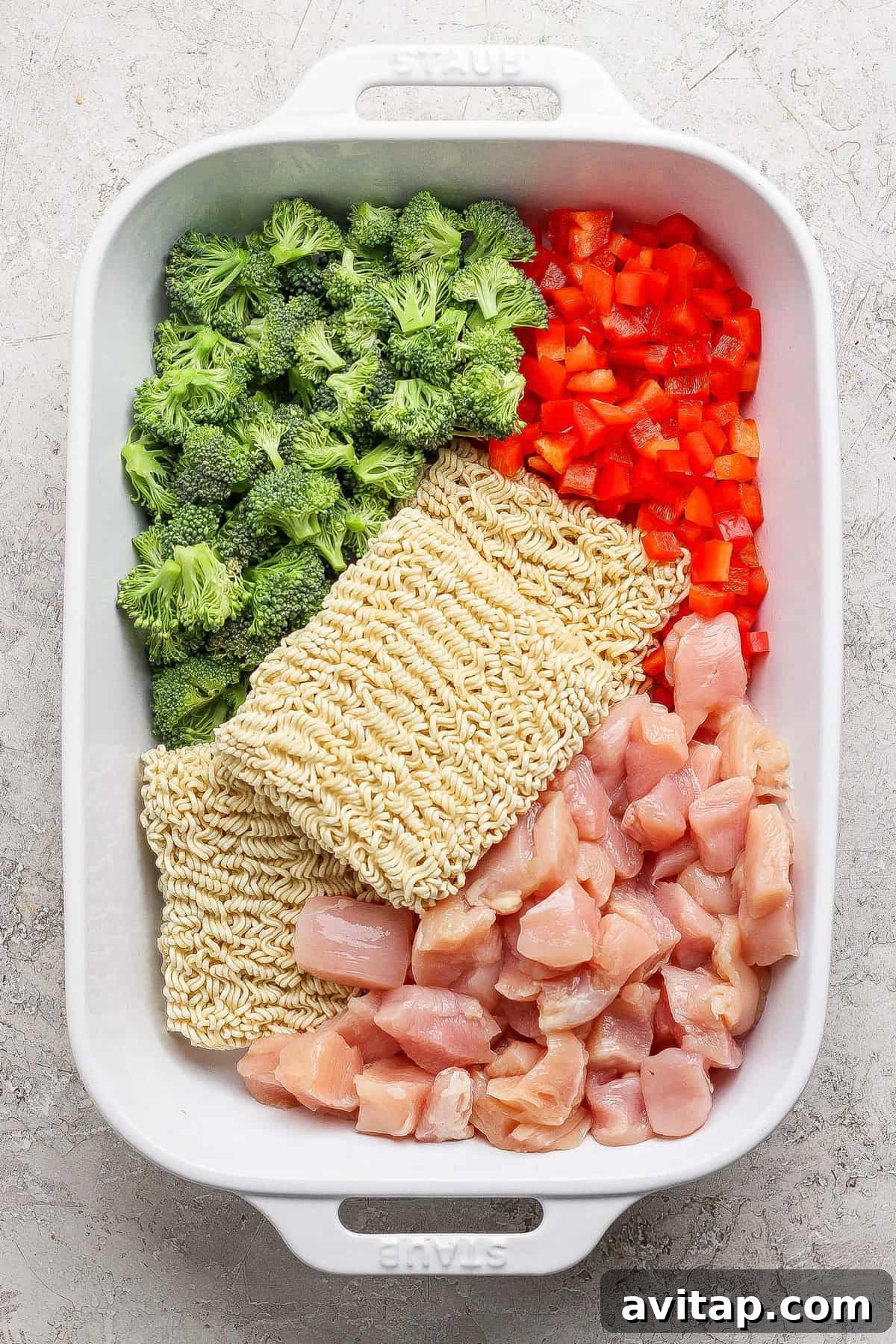 A white baking dish containing raw broccoli florets, diced red bell pepper, two blocks of dry ramen noodles, and chunks of raw chicken, all arranged separately.